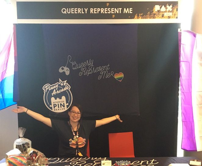 A femme looking person looking excited with their arms, sitting at the Queerly Represent Me booth.