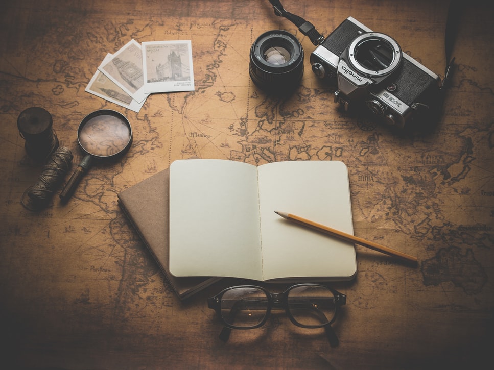 A notebook, reading glasses, a magnifying glass, and old-fashioned camera on top of a monochrome map of the world.